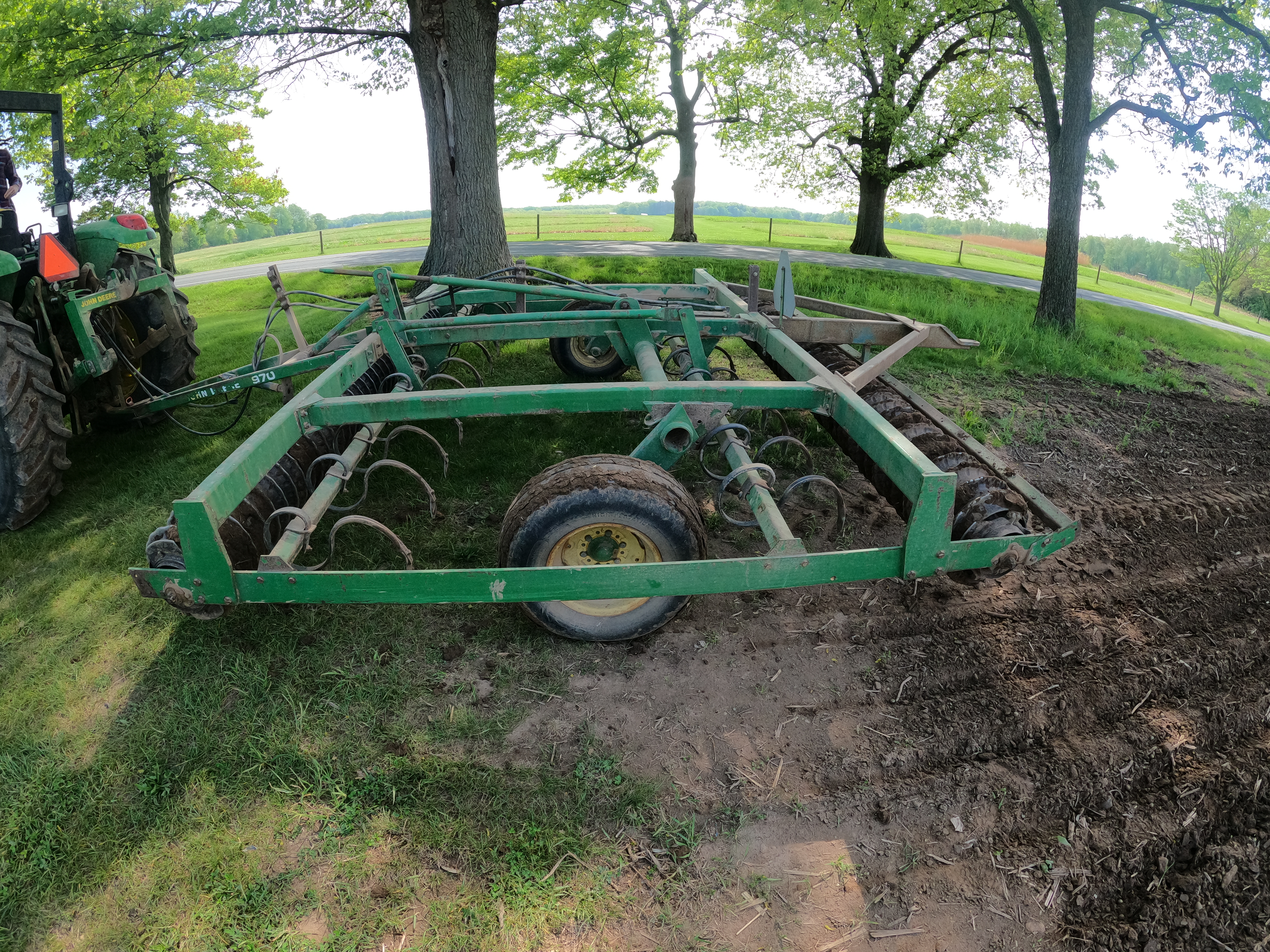A green piece of farm machinery with many hooked teeth being pulled by a green tractor.